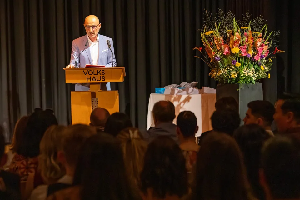 Referent am Volkshaus-Podium vor sitzendem Publikum neben buntem Blumenarrangement